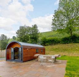 A small arched wooden pod with an open door on a stone patio next to a hot tub and two white chairs and a table at Stiperstones Pod in White Grit near Chirbury