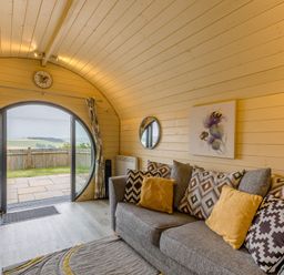 A living room with a gray sofa and patterned cushions near a circular glass door at Stiperstones Pod in White Grit near Chirbury