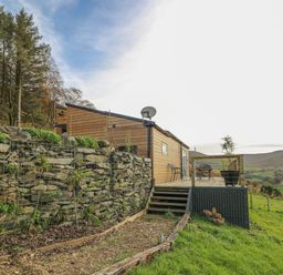 A wooden lodge with an outdoor deck and chairs overlooking a green hillside with trees at Haka Lodge in Devil's Bridge