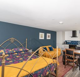 A bedroom with a bed floral bedding a yellow throw a yellow sofa with blue cushions a dining table with chairs and a kitchen area at Haka Lodge in Devil's Bridge