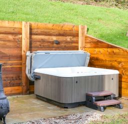 An outdoor hot tub with cover and steps on a stone patio next to a metal chiminea and wooden fence with grass background at Little Silver Fox in High Bickington