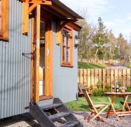 An outdoor area with wooden chairs and a table with wine glasses next to a grey corrugated metal building with wooden framed windows and door at Little Silver Fox in High Bickington