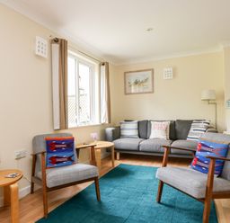 A living room with two chairs, a sofa, a blue rug, wooden side tables, and a window with curtains at Pugwash Cottage in Falmouth