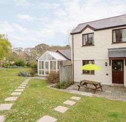 An exterior view of a two-story house with a wooden door a picnic bench with a yellow umbrella on a paved patio and a garden path leading to a glass conservatory at Pugwash Cottage in Falmouth