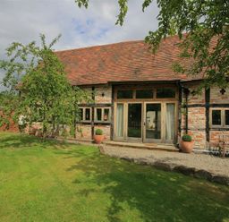 A garden with trees and grass outside a brick and timber building with wooden doors and windows at Whites Farm Barn in Ledbury