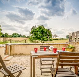 An outdoor patio area with a wooden table and chairs with red cups and a covered dish overlooking a garden and fields at Top Barn in Chipping Norton