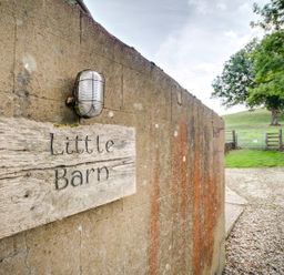 A wooden sign reading Little Barn mounted on a concrete wall next to a gravel driveway with a field and trees in the background at Little Barn Ascott near Chipping Norton