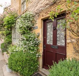 A brown front door with lace curtains surrounded by green plants and flowers on a stone building at Benfield in Stow-on-the-Wold