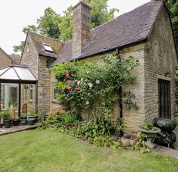 A stone house with a glass conservatory surrounded by potted plants and a garden with flowers and greenery