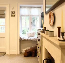 A room with a window seat with cushions a door a chest of drawers a fireplace with candles and plants and a stuffed hippopotamus at Hare House in Chipping Norton