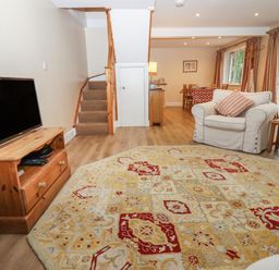 A living room with a patterned rug a television on a wooden stand armchairs and a dining area at The Cottage At Barrow Mead in Bath