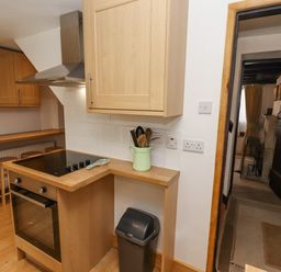 A kitchen with wooden cabinets a black stove a trash bin and a doorway leading to another room at Newbury Cottage in Bourton-On-The-Water