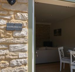 A stone wall with a sign reading saddlebacks and an open door showing a sofa dining table and chairs inside at Saddlebacks Barn Burford