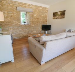 A living room with a beige sofa, wooden coffee table, small TV on a glass stand, white cabinet with a lamp, and stone wall at Saddlebacks Barn in Burford