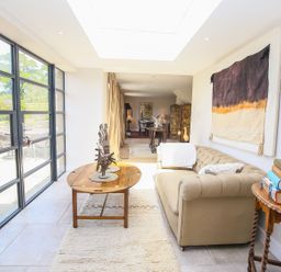 A sitting area with a beige sofa a wooden coffee table with abstract sculptures and a side table with books next to large windows at Fairview Cottage in Milton-Under-Wychwood