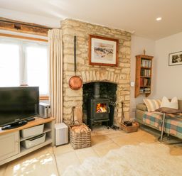 A living room with a stone fireplace wood stove TV on a cabinet and a sofa with cushions at The Lodge in Coln St Aldwyns