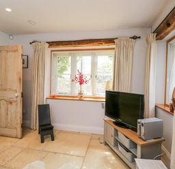 A room with a wooden door a black chair a window with curtains curtain rods and a TV on a wooden cabinet at The Lodge in Coln St Aldwyns