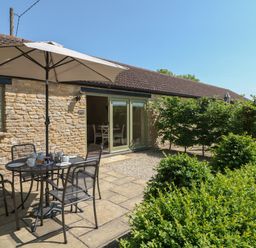 An outdoor patio area with a metal table and chairs under a large umbrella in front of a stone building with green windows and doors at Ewecub Barn in Burford