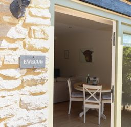A dining area with a round wooden table and white chairs inside a stone building at Ewecub Barn Burford