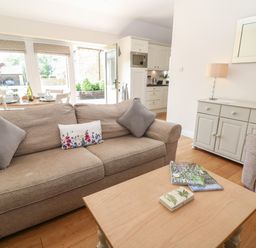 A living room with a beige sofa, a wooden coffee table with magazines, a sideboard with a lamp and plants at Ewecub Barn in Burford