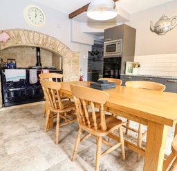 A kitchen with wooden dining table and chairs a black stove under a stone arch clock on wall and modern cabinets at South Hill Farmhouse in Stow On The Wold