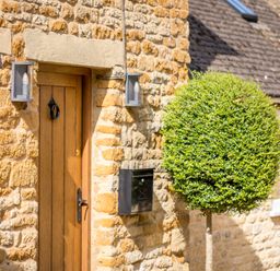 A wooden front door with black handle and knocker on a stone wall with two lights and a black mailbox beside a round trimmed bush at Orchard House in Stow-On-The-Wold