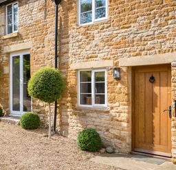 Exterior of a stone house with a wooden door and white framed windows with spherical and bushy plants at Orchard House in Stow-On-The-Wold