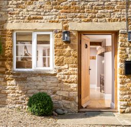 A stone exterior wall with a wooden door open to a hallway and two trimmed round bushes at Orchard House in Stow-On-The-Wold