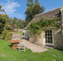 An exterior view of a stone cottage with a small garden table and chairs on grass at Mayfly Cottage Coln St Aldwyns near Bibury