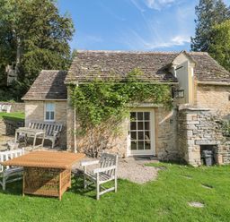 An outdoor seating area with wooden chairs and a table in front of a stone cottage with a tiled roof and greenery at Mayfly Cottage Coln St Aldwyns near Bibury