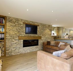 A living room with two sofas a stone wall with a mounted TV and fireplace and a kitchen area at Old Groom's Cottage in Minster Lovell