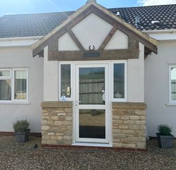 Front entrance of a single story house with a glass door and two windows on either side at Old Groom's Cottage in Minster Lovell