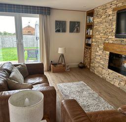 A living room with leather sofas a stone fireplace a wall-mounted TV and glass doors leading to a garden at Old Groom's Cottage in Minster Lovell