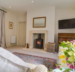 A living room with a wood stove fireplace a television on the wall and flowers on a table at The Old Dairy in Shilton