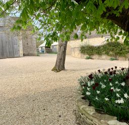An outdoor courtyard with a large tree, flower bed, gravel ground, and stone buildings at The Old Dairy in Shilton