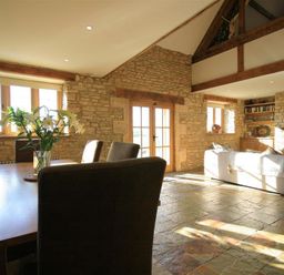A living and dining area with a wooden table and chairs a sofa and stone walls at Calcot Peak Barn Bibury near Northleach