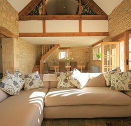 A living room with a beige sectional sofa and floral cushions next to wooden stairs and a dining table with chairs at Calcot Peak Barn Bibury near Northleach