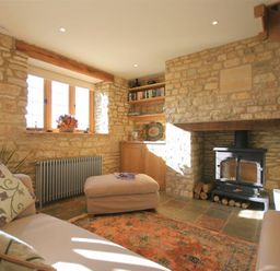 A living room with stone walls a wood stove a beige sofa and an ottoman at Calcot Peak Barn Bibury near Northleach