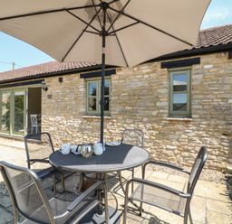 A stone exterior wall with two windows and a glass door opening to a patio with a metal table and four chairs under a large umbrella at Nutchat Barn in Burford