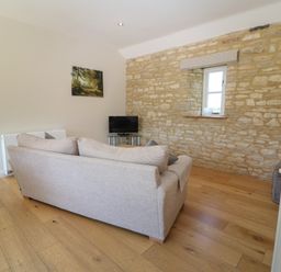 A living room with a beige sofa wooden floor stone wall window a small TV on a stand and a framed picture on the wall at Nutchat Barn in Burford