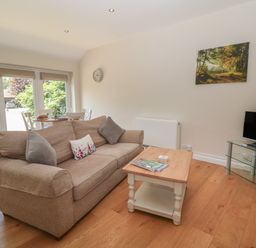 A living room with a beige sofa a wooden coffee table a wall clock a television on a glass stand and a dining table near glass doors at Nutchat Barn in Burford