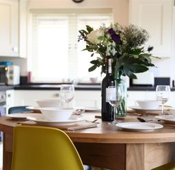 A kitchen with a wooden dining table set with white plates bowls wine glasses a bottle of wine and a vase of flowers at Park Bothy Wyck Rissington