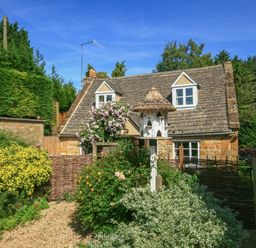 A stone cottage with two dormer windows surrounded by a garden with flowers and a birdhouse at Hadcroft Cottage in Aston Magna