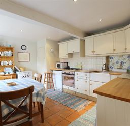 A kitchen with a wooden dining table covered with a checkered tablecloth and wooden chairs a cabinet with dishes a counter with a stove microwave and sink and striped rugs on the floor at The Mews Cottage in Tisbury