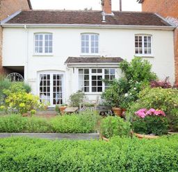 A garden with various plants and flowers in front of a white two-story house with multiple windows and a wooden bench at The Mews Cottage in Tisbury