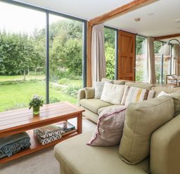 A living room with a green sofa and wooden coffee table by large glass windows showing a garden at The Cart Barn in Ridge near Chilmark