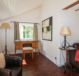 A room with a wooden desk and stool in front of a window a black leather chair a small round table with a lamp and a sofa with cushions and a blanket at Gun Hill Cottage in Heathfield