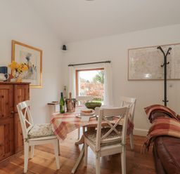 A dining area with a wooden table covered with a checked tablecloth surrounded by four white chairs with cushions a wooden cabinet with a pitcher and flowers a framed picture on the wall a coat rack a brown leather sofa with a blanket and a window showing greenery at Russet in Martin