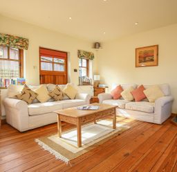 A living room with two sofas, a wooden coffee table, wooden floors, and windows with curtains at The Coach House in Lympstone