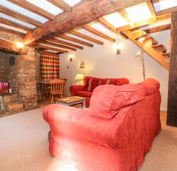 A living room with red sofas a wooden dining table and chairs a stone fireplace and exposed wooden beams at Spring Cottage in Wroxton